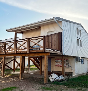 Maison sur pilotis avec terrasse bois : Location saisonnière. Cabane sur pilotis, bardage blanc, grande terrasse bois avec garde-corps croisés. Affiche publicitaire sur la base.