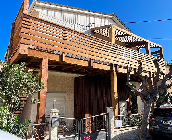 Terrasse en bois sur pilotis : l'extension idéale. Façade d'une maison au numéro 17 avec une grande terrasse en bois sur pilotis. Balustrade à lattes horizontales et petite pergola attenante.