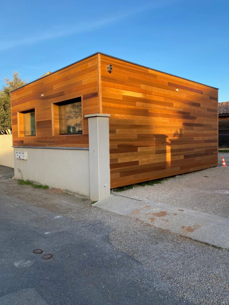 Bureau de jardin moderne en bois avec bardage naturel Petit bâtiment moderne avec bardage bois naturel, socle clair et fenêtres carrées sous un ciel bleu azur.