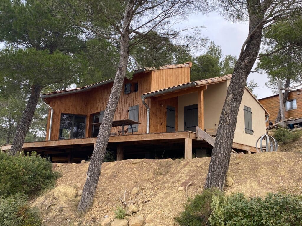 Chalet moderne en bois avec grande terrasse sur pilotis, niché dans une pinède escarpée sous un ciel voilé.