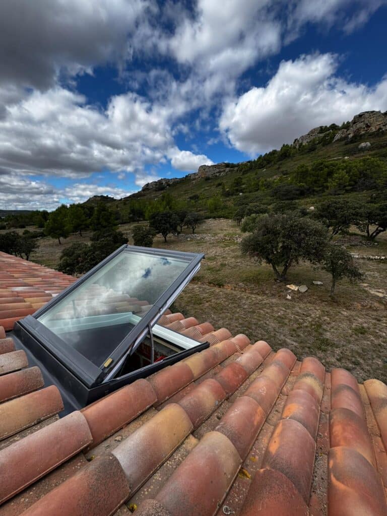 Fenêtre de toit ouverte avec vue sur collines verdoyantes Fenêtre de toit ouverte sur tuiles romanes avec vue panoramique sur collines boisées et ciel nuageux.