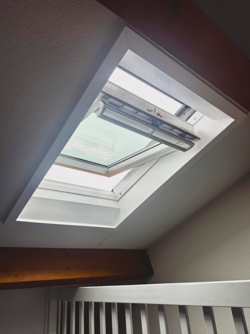 Fenêtre de toit type Velux ouverte dans une pièce mansardée avec poutres apparentes apportant de la lumière naturelle.